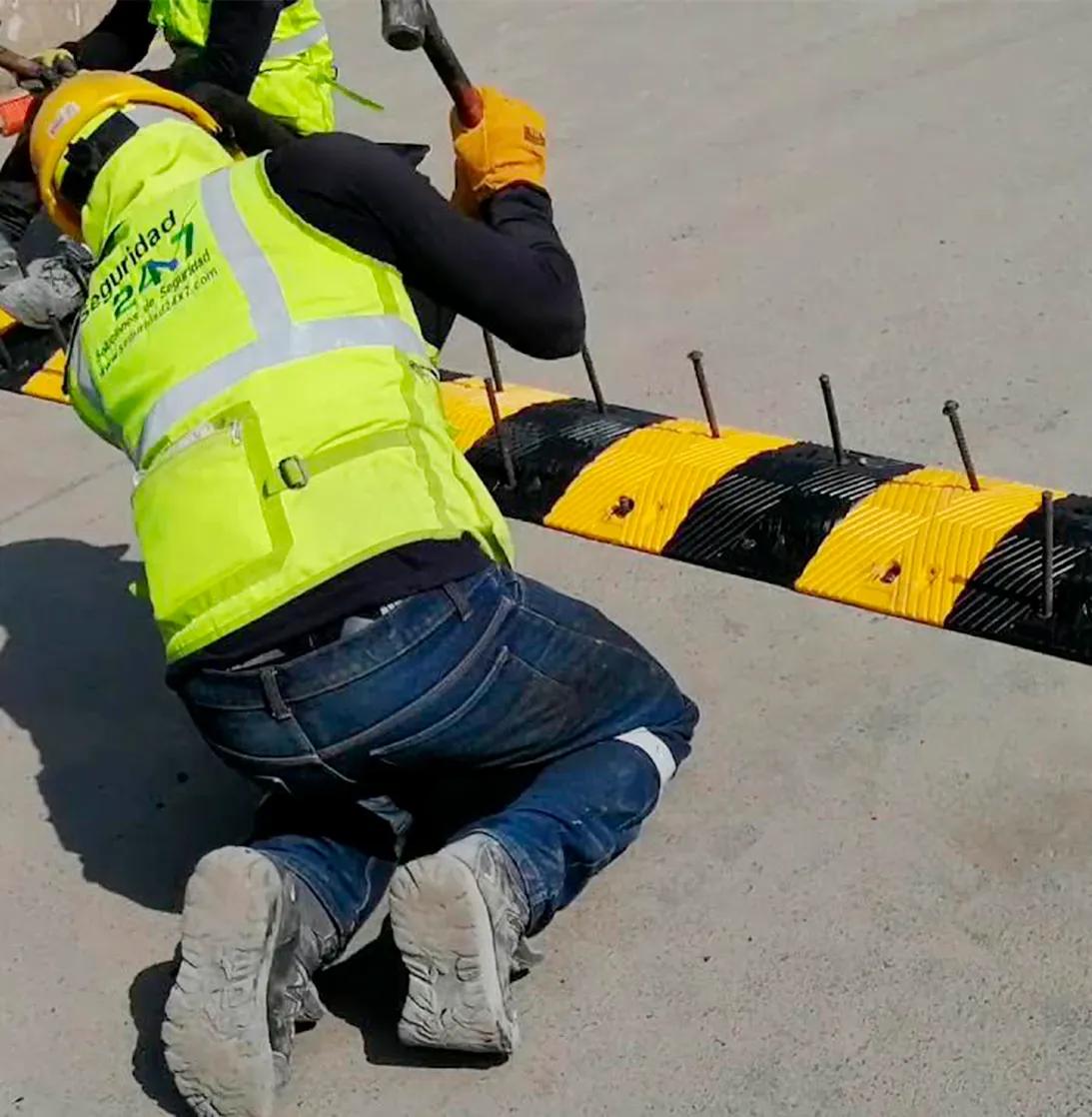 Trabajador con casco y chaleco fluorescente fija con clavos un tope de velocidad amarillo y negro sobre concreto