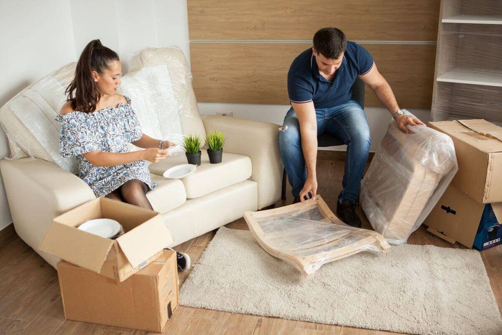 Pareja empaca una sala: sofá cubierto con plástico, silla envuelta en película y cajas de cartón alrededor