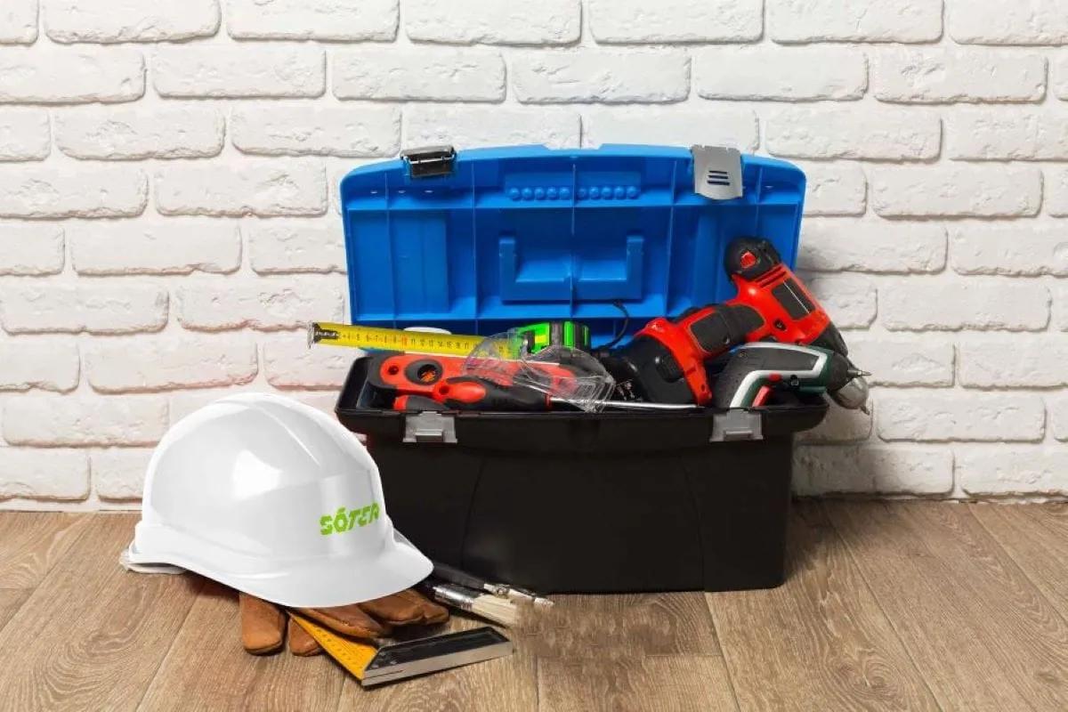 Casco de seguridad blanco con logo verde junto a una caja de herramientas azul con taladro, flexómetro y guantes