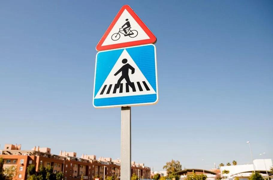 Señal triangular de ciclistas sobre una señal azul de paso peatonal en un poste, con cielo despejado