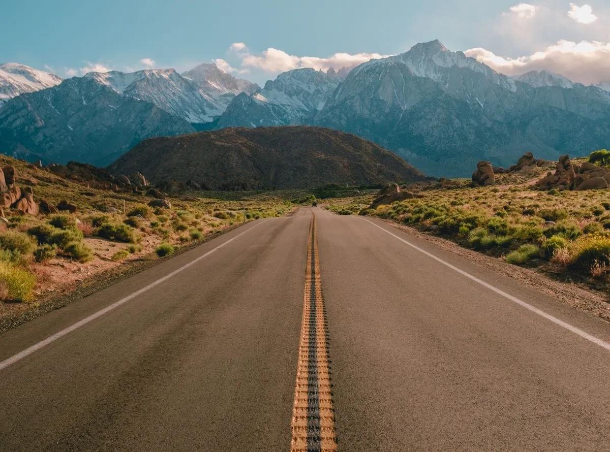 Carretera recta de dos carriles con doble línea amarilla que lleva a montañas nevadas, de día