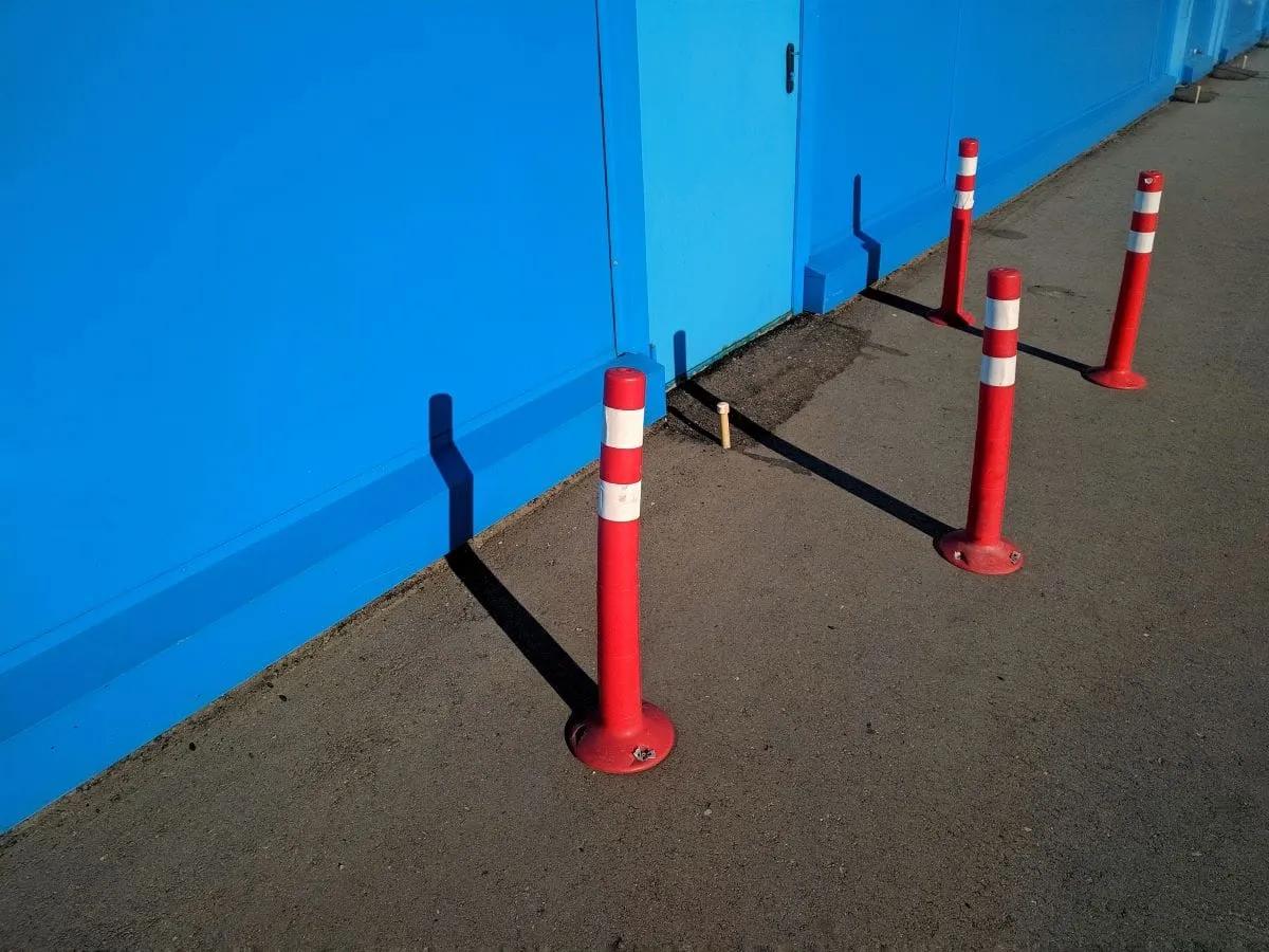 Postes delineadores flexibles rojos con bandas blancas reflejantes junto a un muro azul