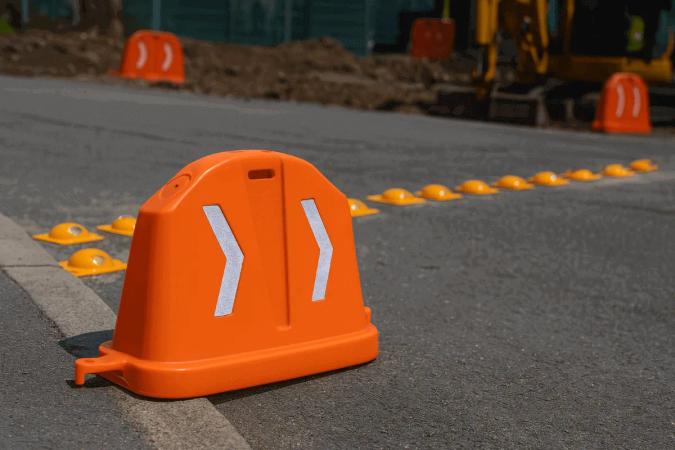 Barrera anidable naranja con dos chevrons blancos junto a una hilera de boyas amarillas en una calle en obra
