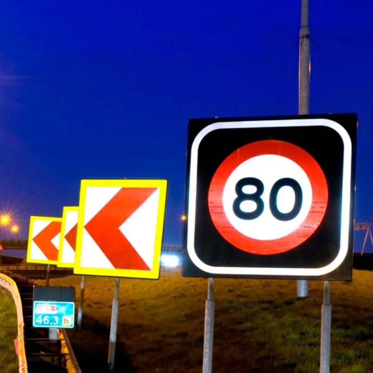Señal de límite de 80 y tableros de curva con flechas rojas reflejando la luz junto a una carretera de noche