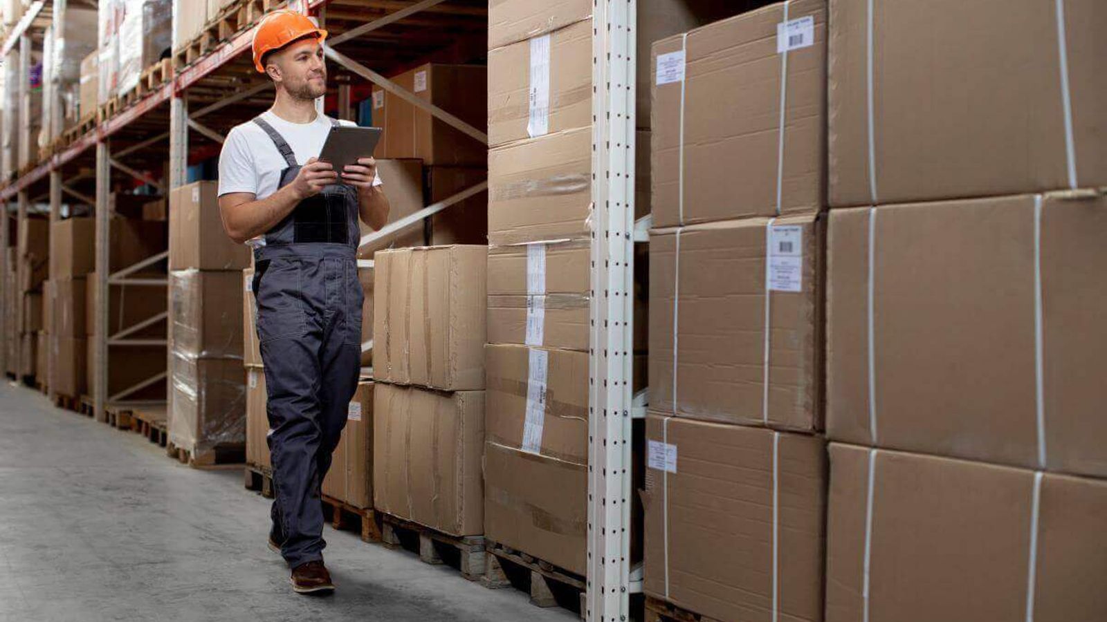 Trabajador con casco y tableta recorre un almacén entre tarimas de cajas de cartón flejadas