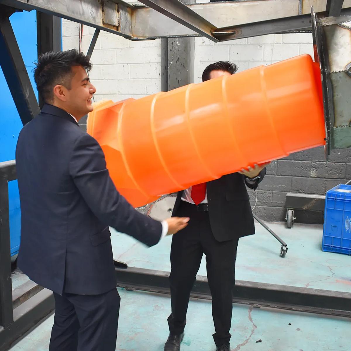 Dos hombres de traje sostienen el cuerpo de un trafitambo naranja junto a un molde en una planta de fabricación