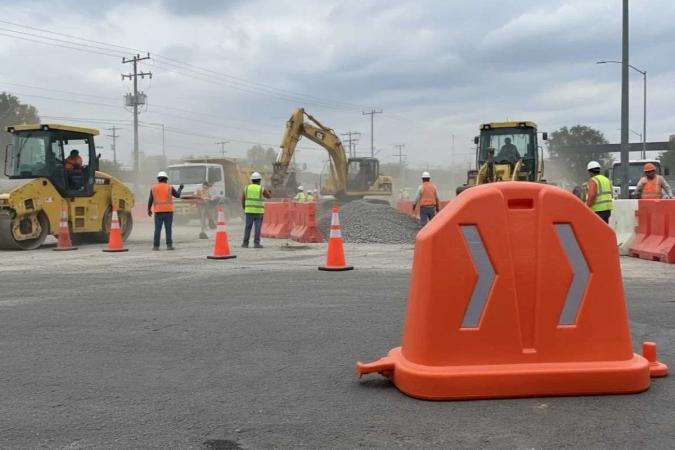 Barrera anidable naranja con flechas reflejantes en una obra, con maquinaria, conos y trabajadores al fondo