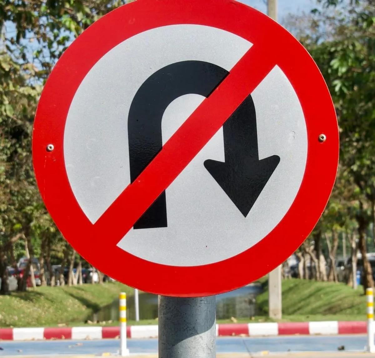 Señal restrictiva circular de prohibido dar vuelta en U, con aro y diagonal rojos, junto a una avenida arbolada
