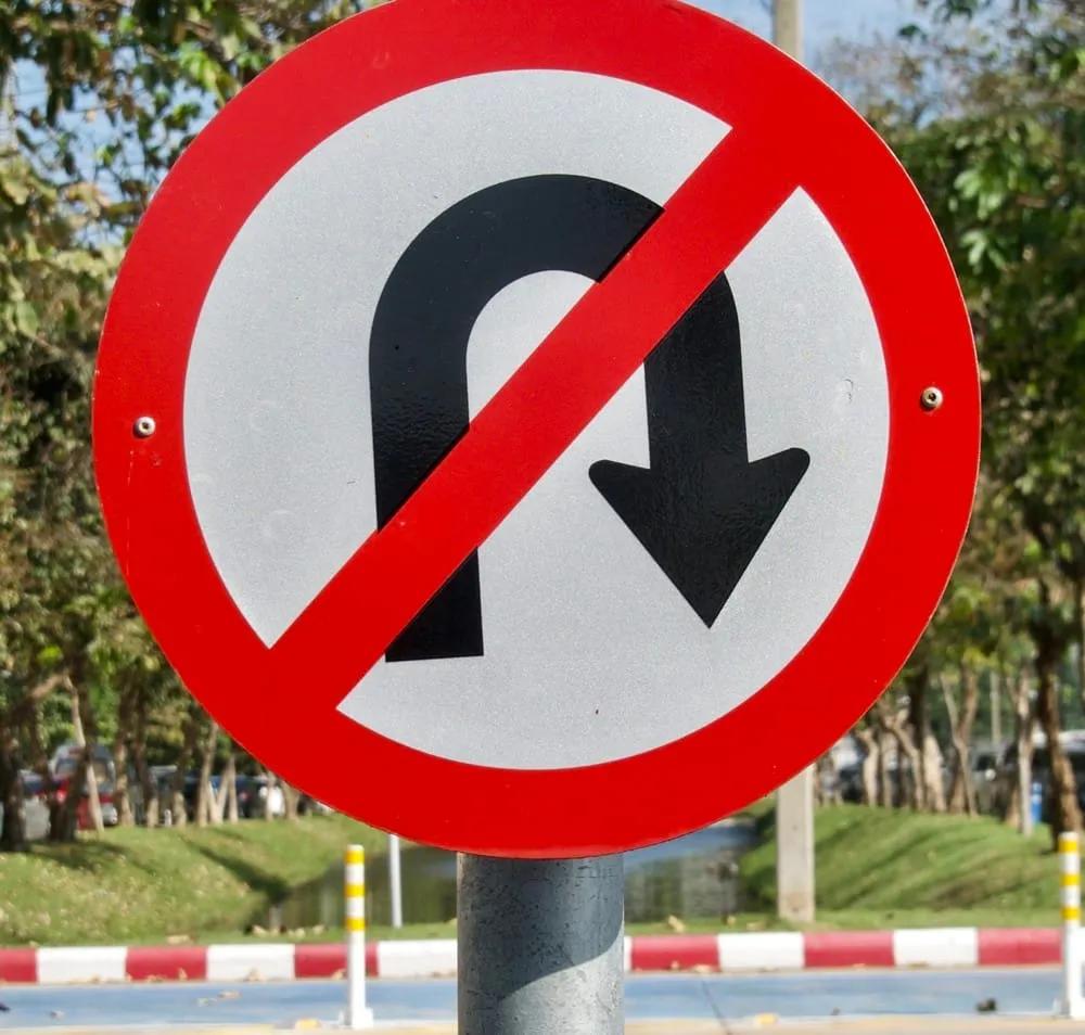 Señal restrictiva circular de prohibido dar vuelta en U, con aro y diagonal rojos, junto a una avenida arbolada
