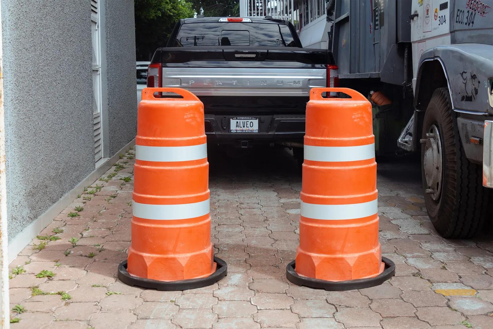 Dos trafitambos naranjas con bandas reflejantes sobre bases negras, en un patio empedrado frente a una pickup y un camión