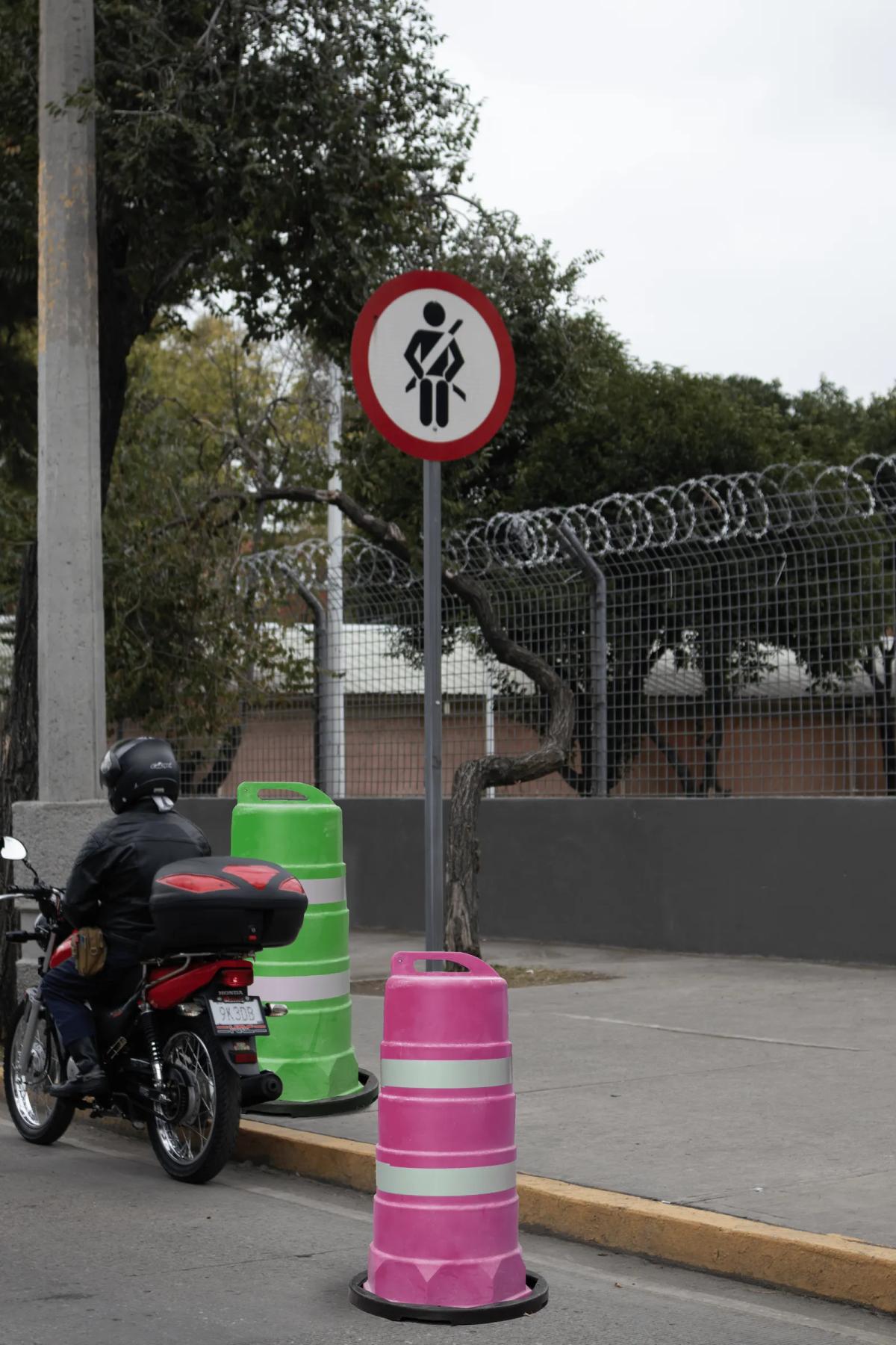 Trafitambos verde y rosa con bandas reflejantes junto a la banqueta, un motociclista y una señal de cinturón de seguridad