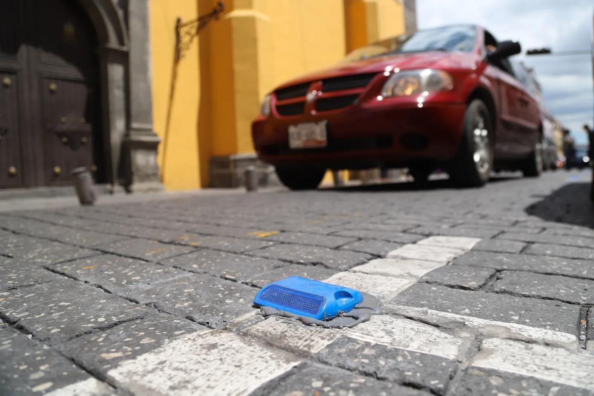 Vialeta reflejante azul fijada sobre una franja blanca en una calle empedrada, con un auto rojo al fondo