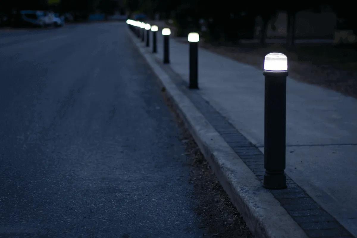 Bolardos negros con luz LED blanca encendida a lo largo de la orilla de una banqueta junto a una calle de noche