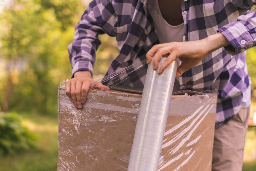 Persona con camisa de cuadros envuelve una caja de cartón con un rollo de playo al aire libre