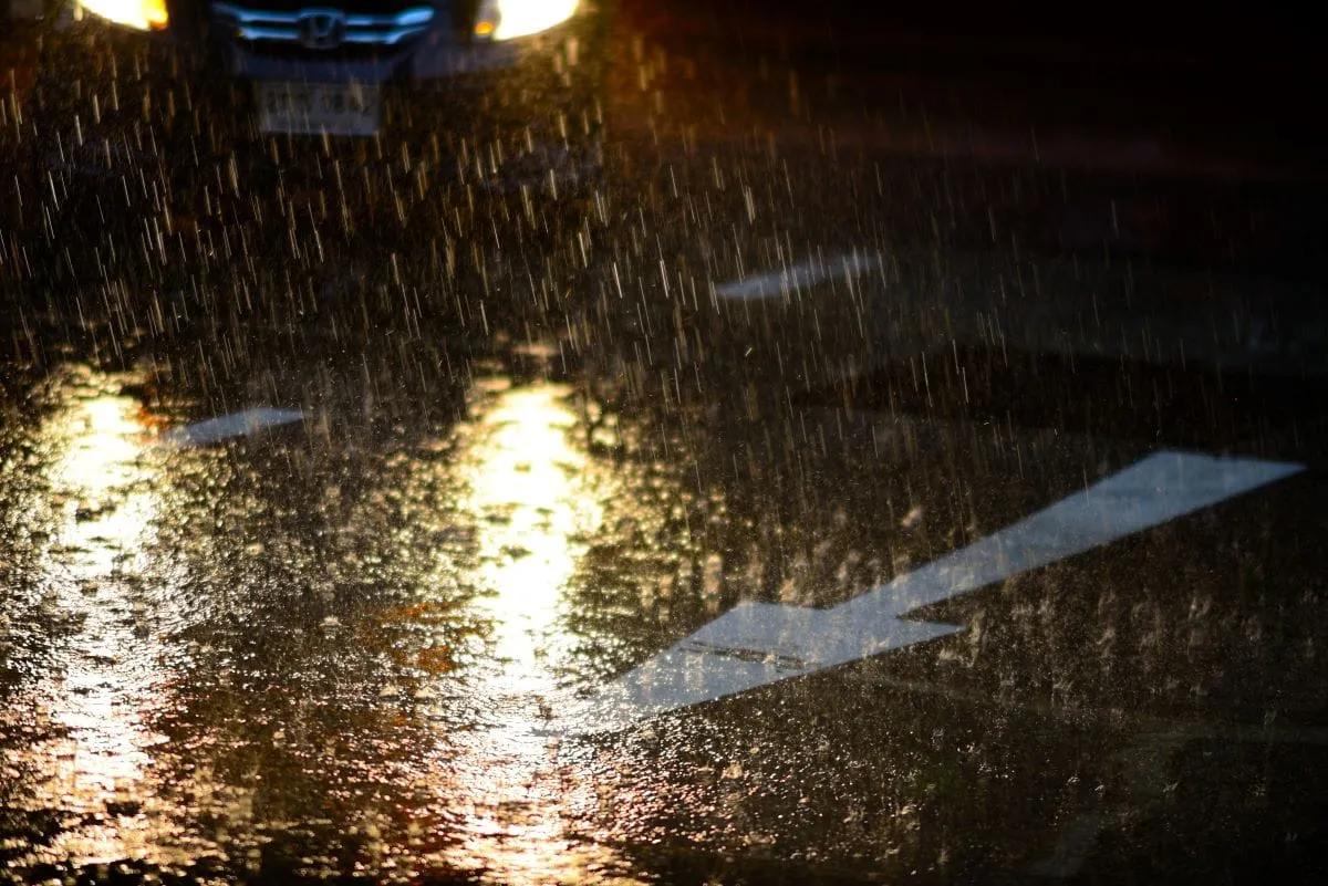 Lluvia intensa de noche sobre el asfalto con una flecha blanca pintada y los faros de un auto que se acerca