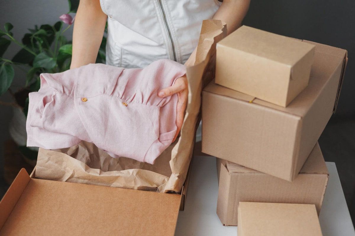 Mujer coloca una blusa rosa en una caja de cartón forrada con papel kraft, junto a cajas kraft apiladas