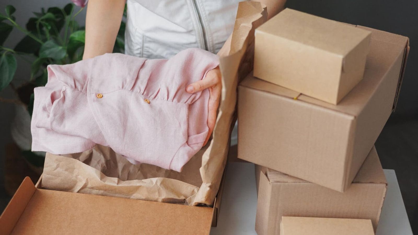 Mujer coloca una blusa rosa en una caja de cartón forrada con papel kraft, junto a cajas kraft apiladas