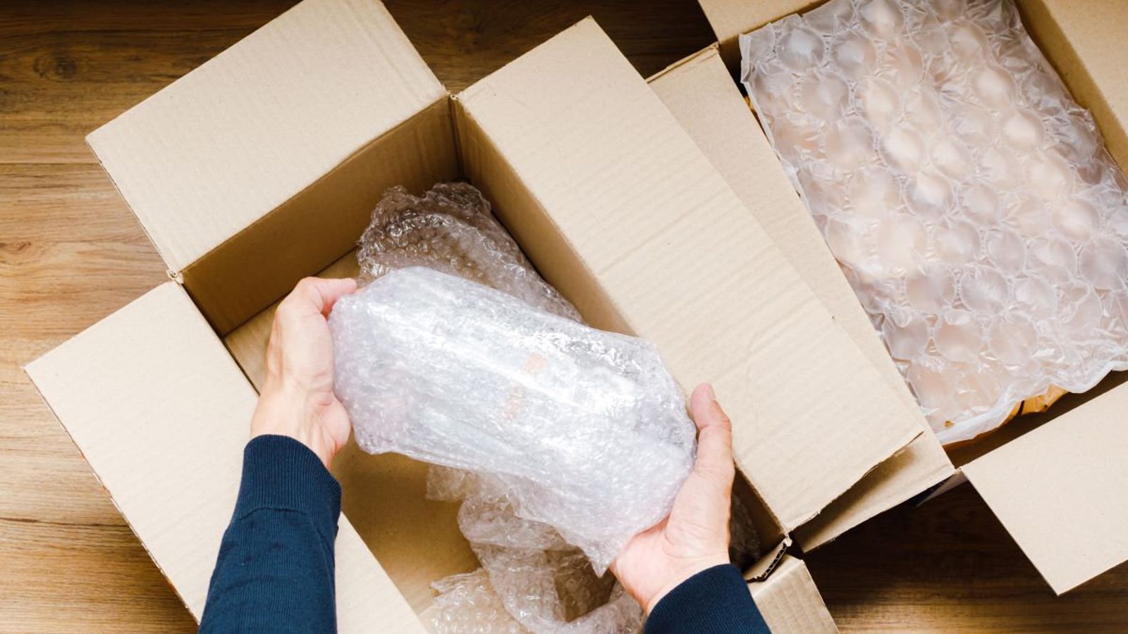 Manos sacan un objeto envuelto en poliburbuja de una caja de cartón abierta, junto a otra caja con más plástico de burbujas