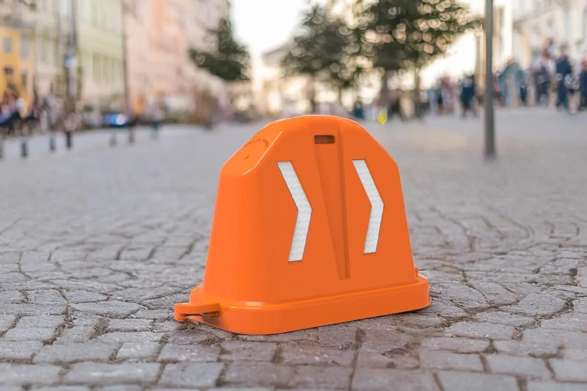 Barrera vial anidable naranja con dos chevrones reflejantes blancos sobre una calle peatonal empedrada con gente al fondo