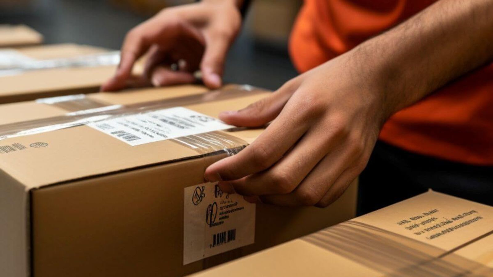 Persona con playera naranja pega etiquetas con código de barras en cajas de cartón selladas con cinta