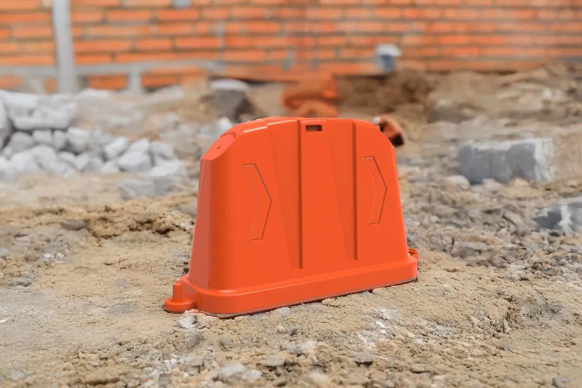 Barrera anidable naranja con flechas en bajo relieve sobre tierra y escombro, frente a un muro de ladrillo