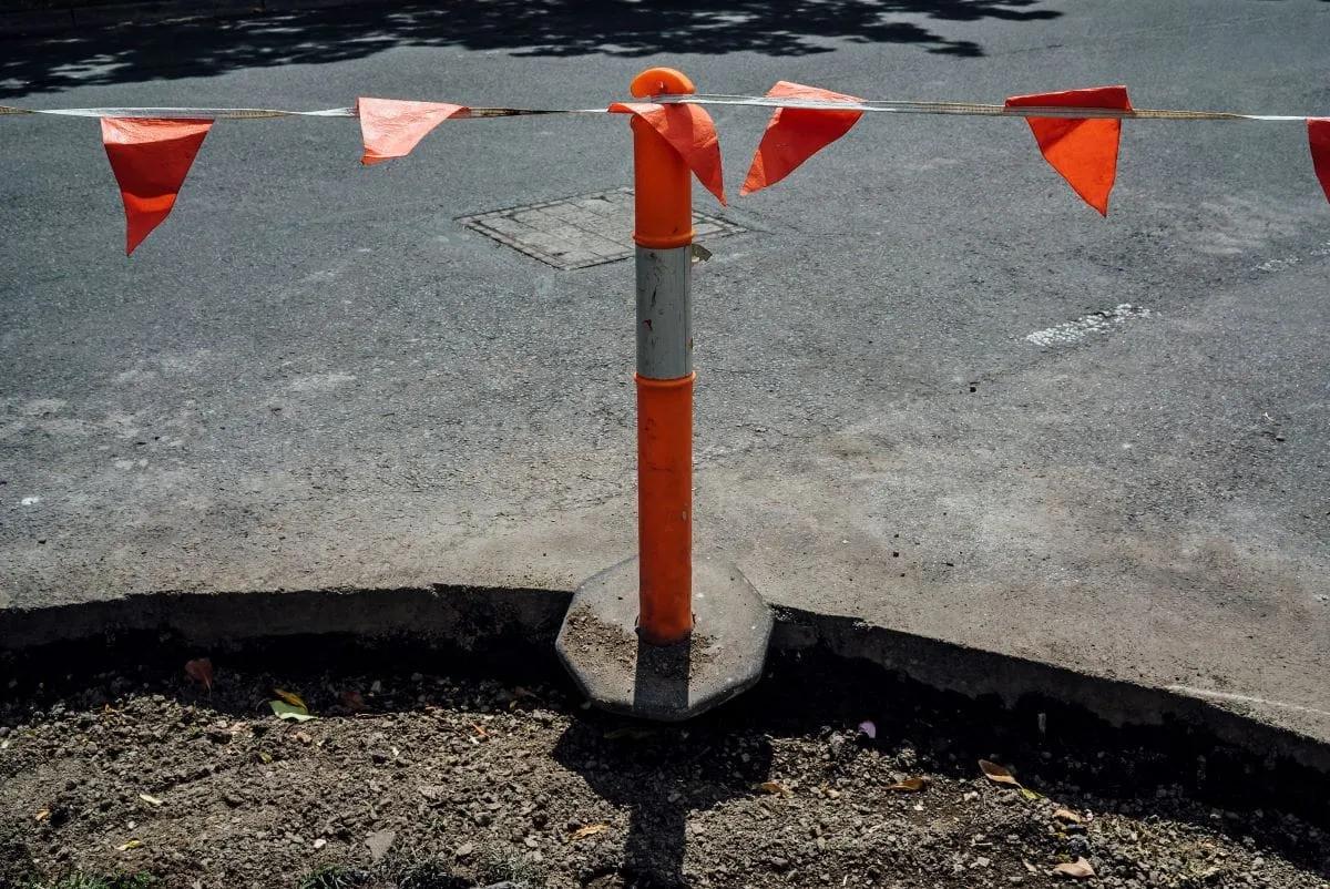 Poste delineador naranja con banda reflejante sostiene una cinta de banderines triangulares naranjas junto a la banqueta