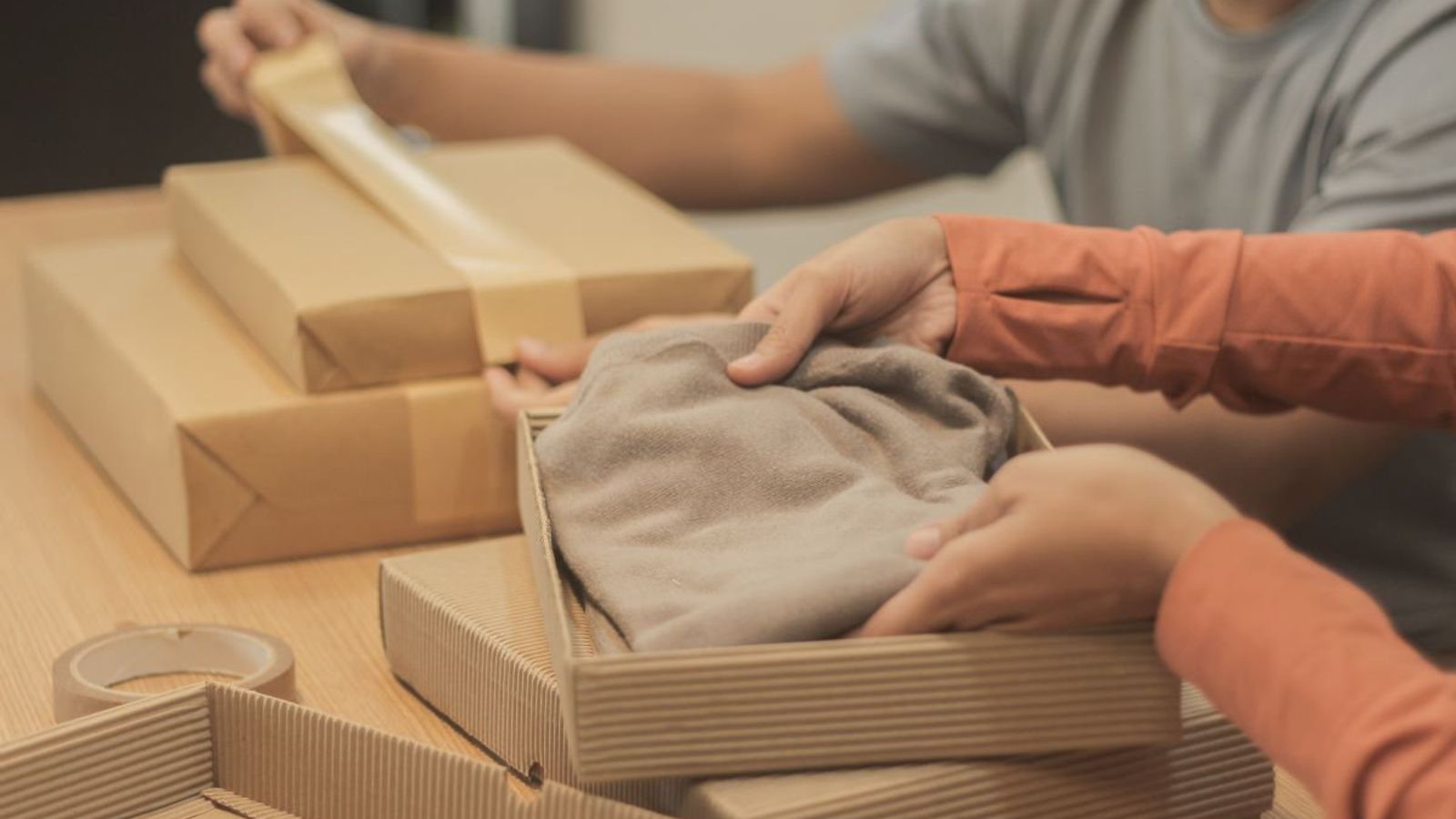 Dos personas acomodan una prenda doblada en una caja de cartón corrugado junto a paquetes envueltos en papel kraft