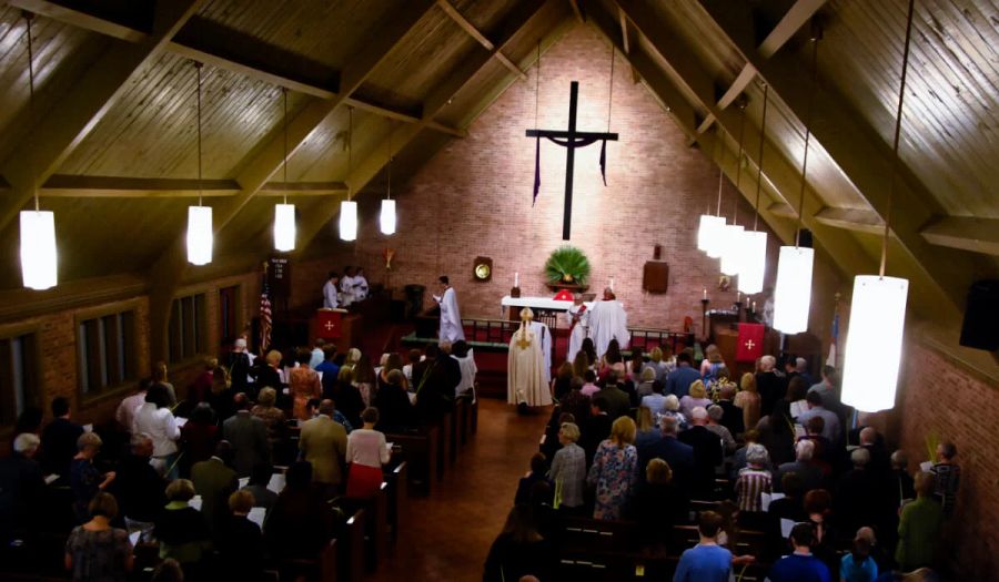 Congregation standing in a church sanctuary during a Palm Sunday service, with clergy in white robes at the altar beneath a large wooden cross and palm branches displayed.