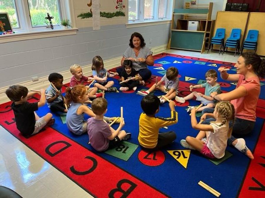 Group of young children sitting in a circle on an alphabet rug in a classroom at St. Francis Episcopal Day School, playing rhythm sticks with two adult teachers.