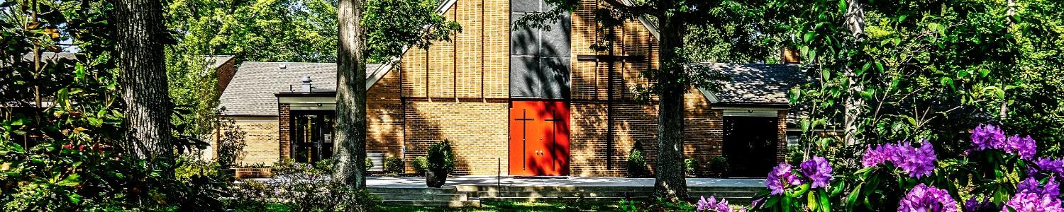Exterior view of a brick church building surrounded by trees and greenery, featuring red front doors with a cross and flowering plants in the foreground.