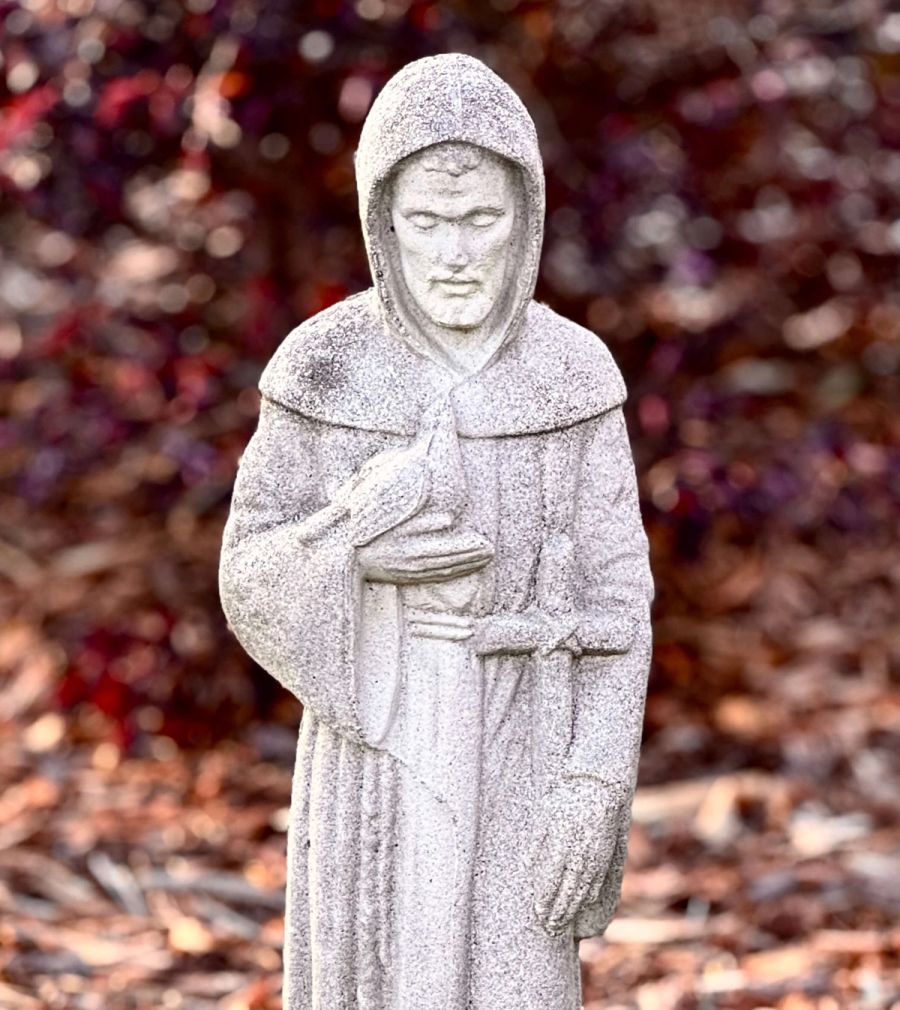A stone statue of a cloaked figure with hands clasped, set against a blurred background of colorful foliage.