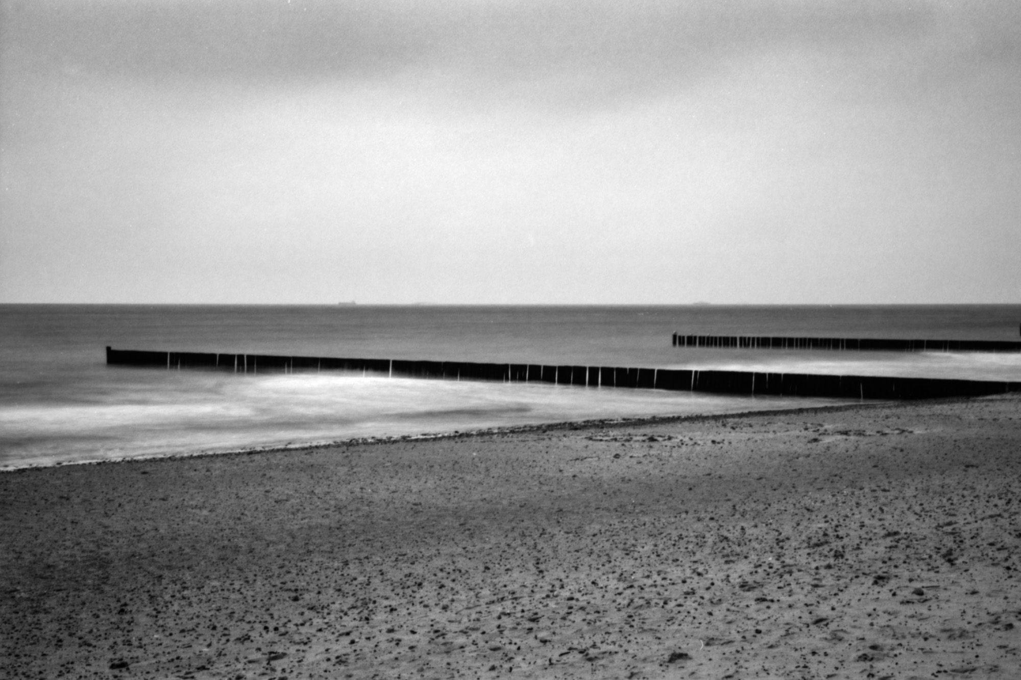 B&W image of a quiet beach with gentle waves softly blurred in a long exposure and piers extending into the sea.