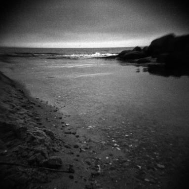 Aufgenommen mit der HOLGA 120 CFN und einem Rotfilter. Stimmungsvoller Strand mit Wellen und Steinen und einer starken Vignette.