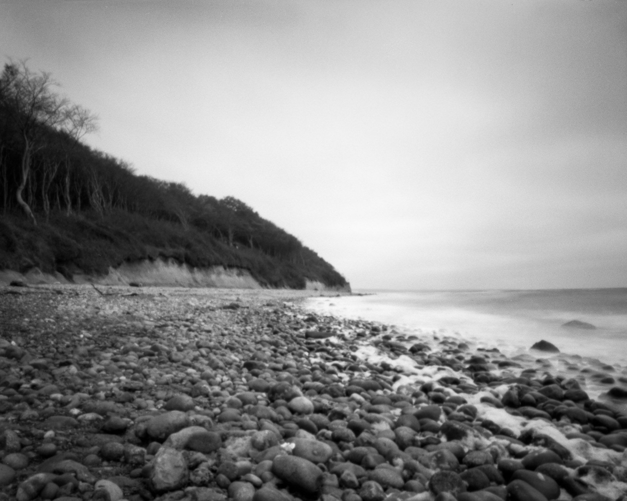 Black and white picture taken with a pinhole camera. Rough rocks are scattered along the edge of the beach with a view of the steep coast and the sea.