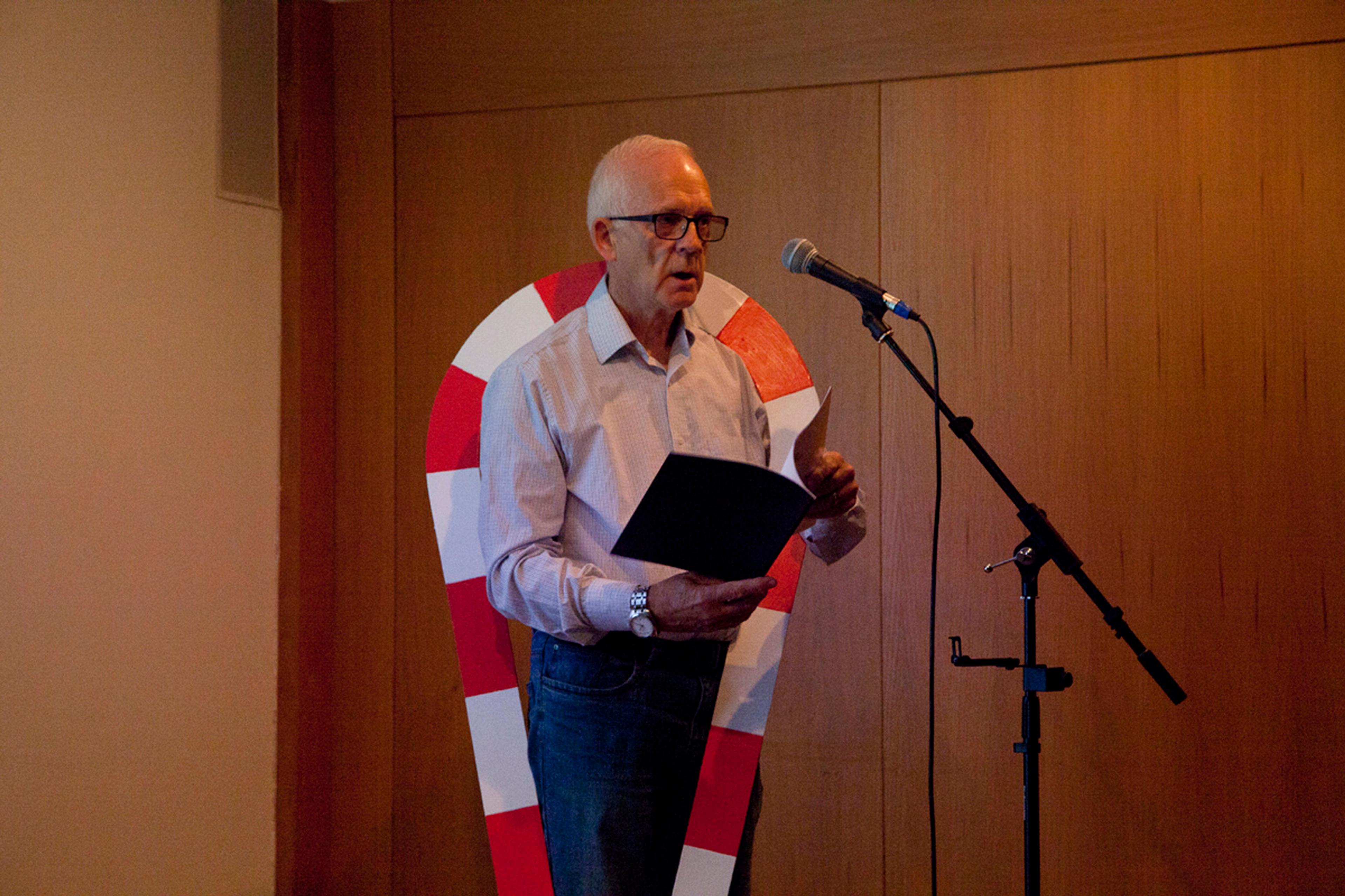 Fabian Peake reading into a microphone, a sculpture hangs over his shoulders