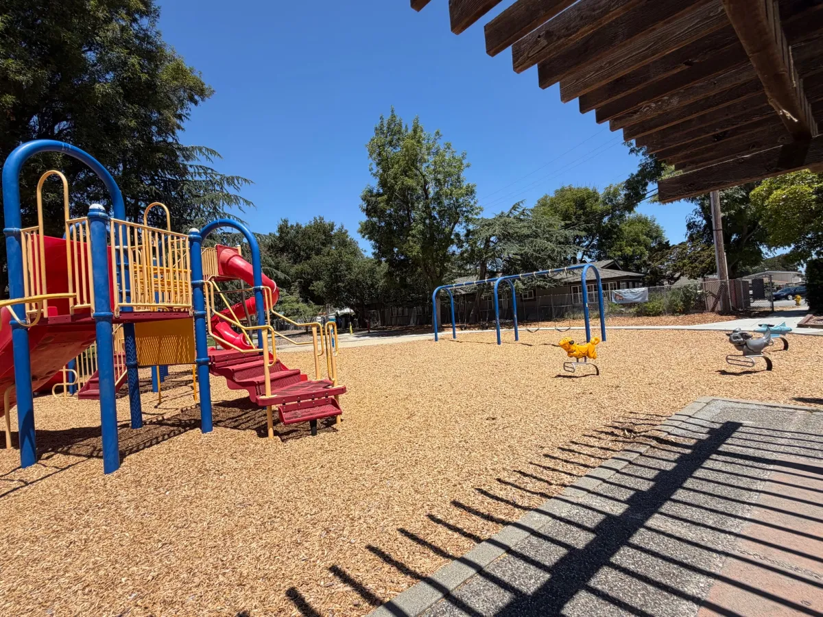 View from picnic tables of swings, ride on toys, and playstructure