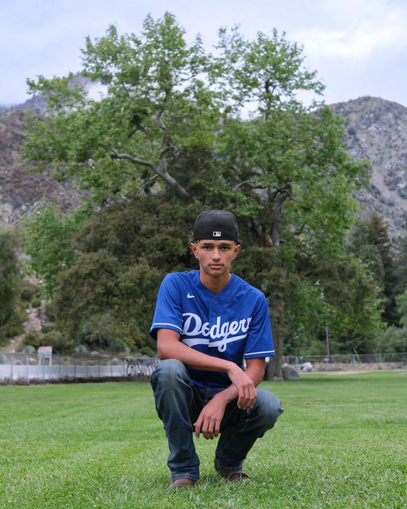 Teen in a Dodgers jersey crouching on the grass under a big tree