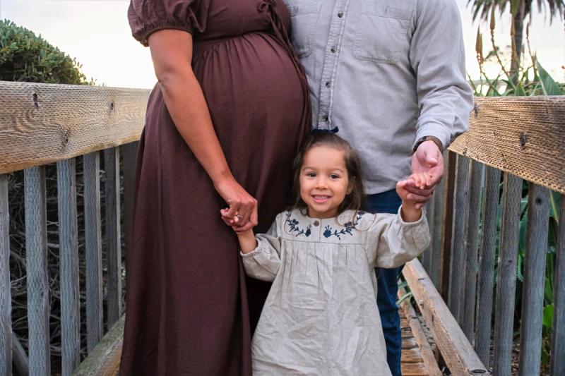 Toddler holding her expecting parents’ hands on a wooden bridge
