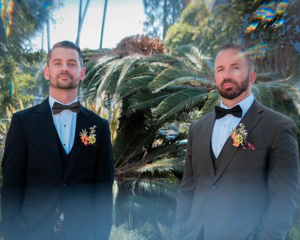 Two grooms with boutonnieres smiling among tropical palms