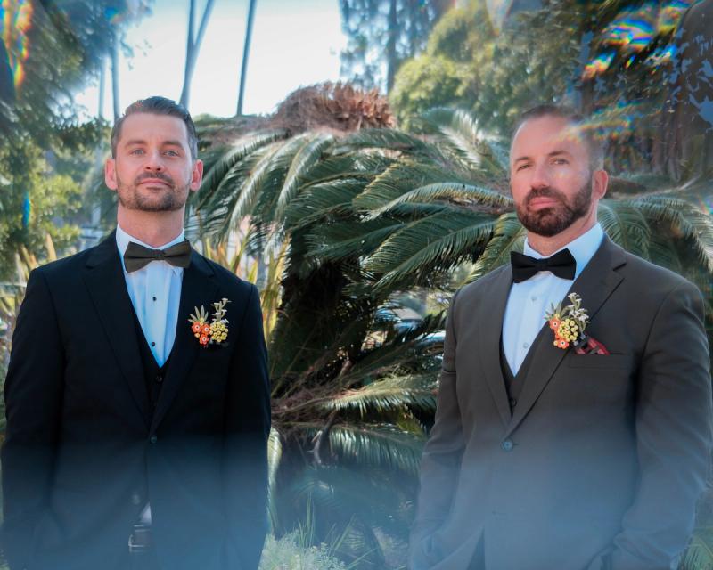 Two grooms with boutonnieres smiling among tropical palms