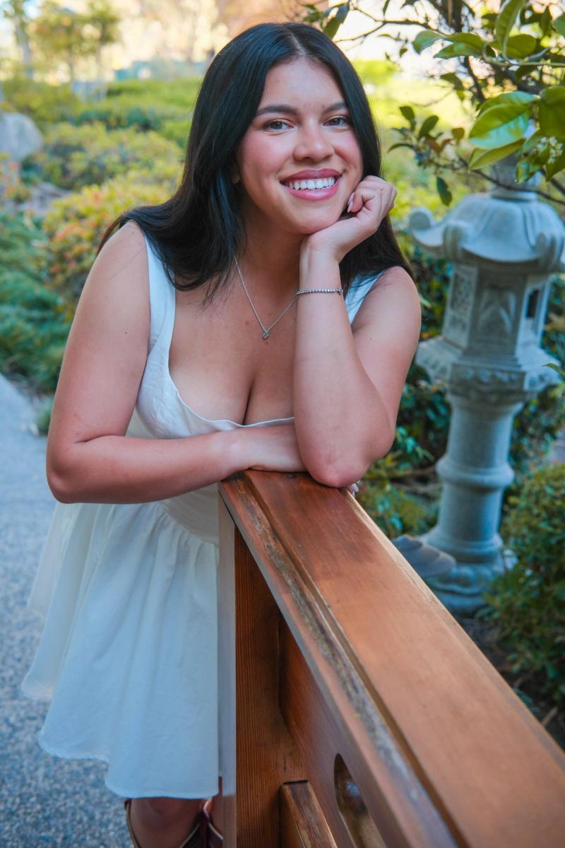 Graduate in a white dress leaning on a wooden railing, chin in hand