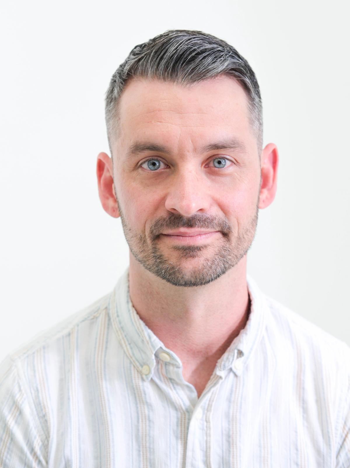 Close studio headshot of a man in a light striped shirt, looking directly at the camera