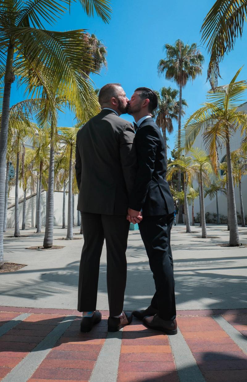 Grooms kissing on a palm-lined walkway under a blue sky