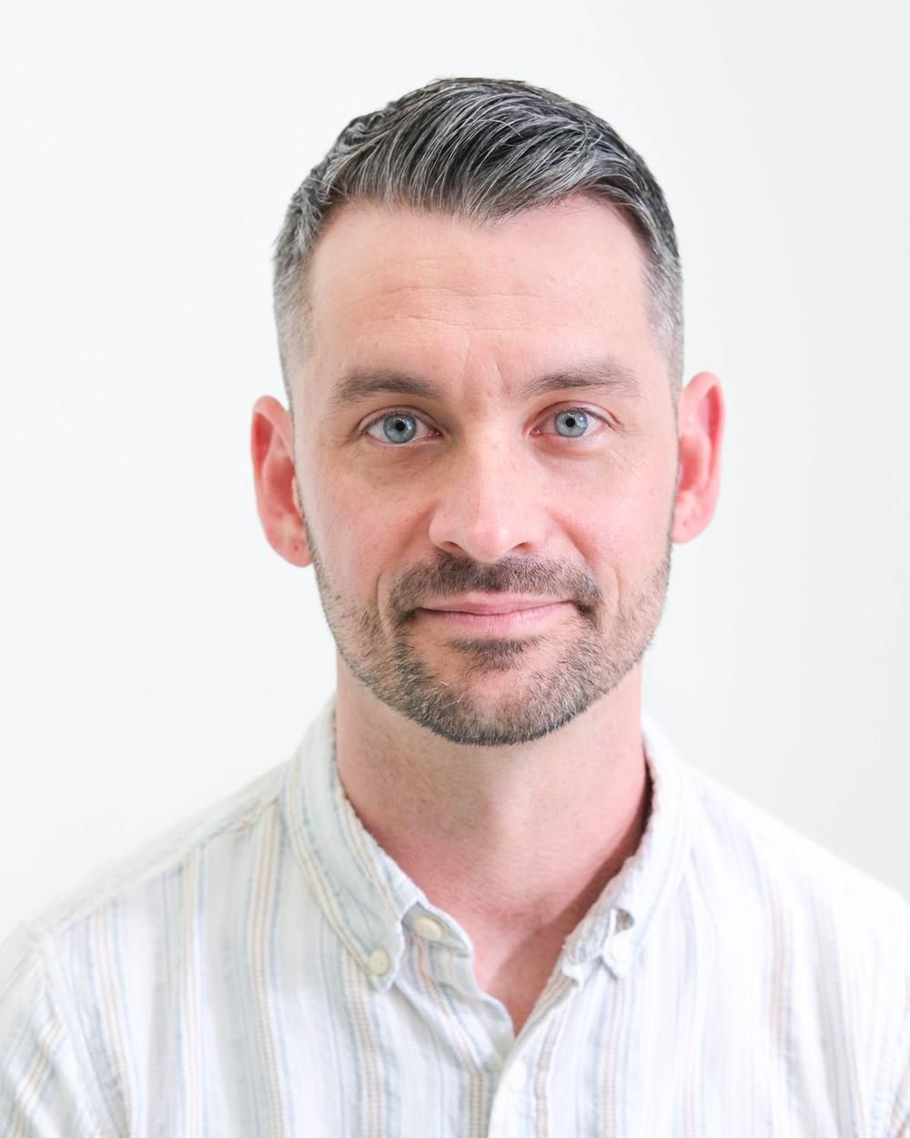 Professional studio headshot of a man in a light button-down shirt, looking directly at the camera