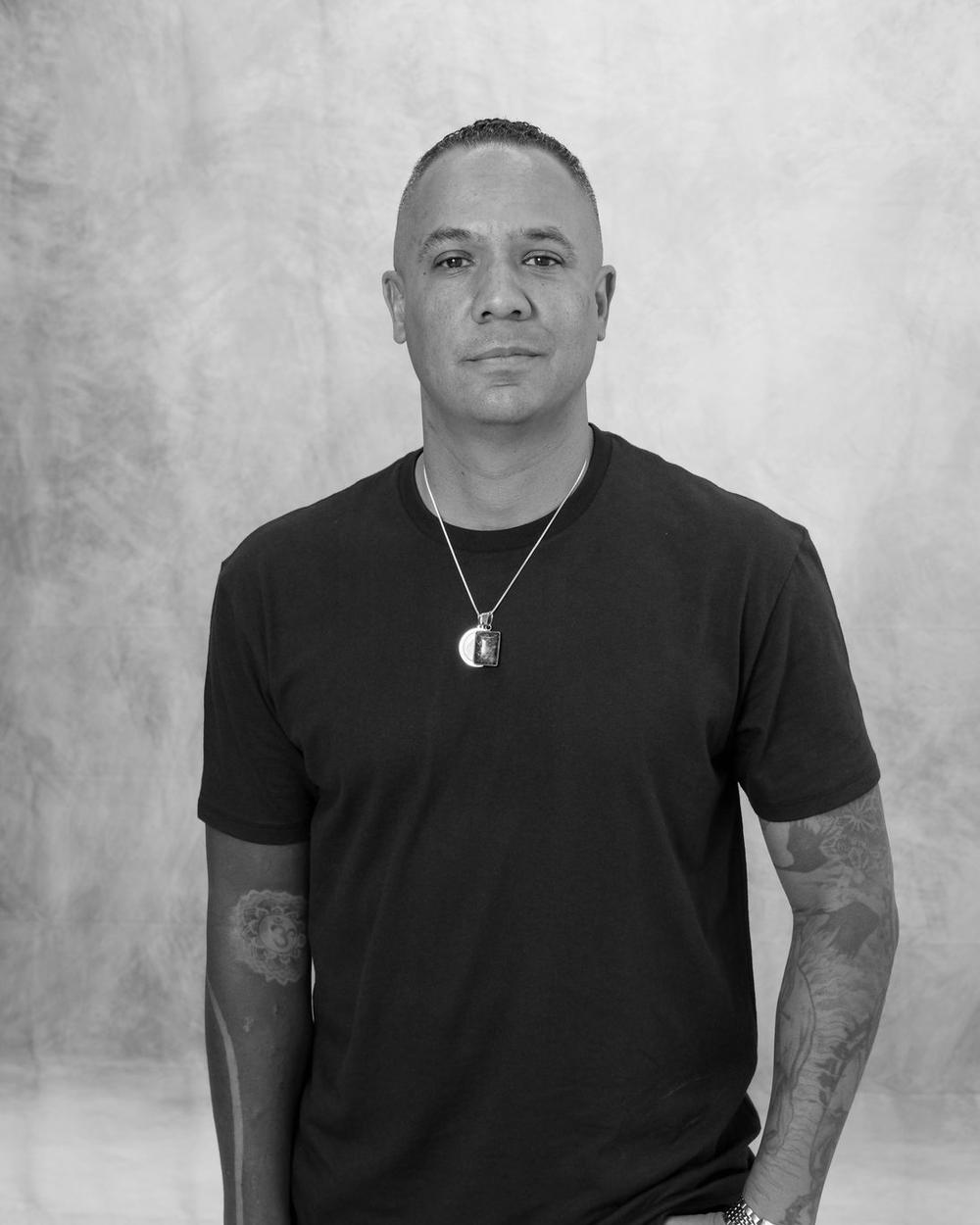 Black and white studio portrait of a man in a black tee against a painted canvas backdrop