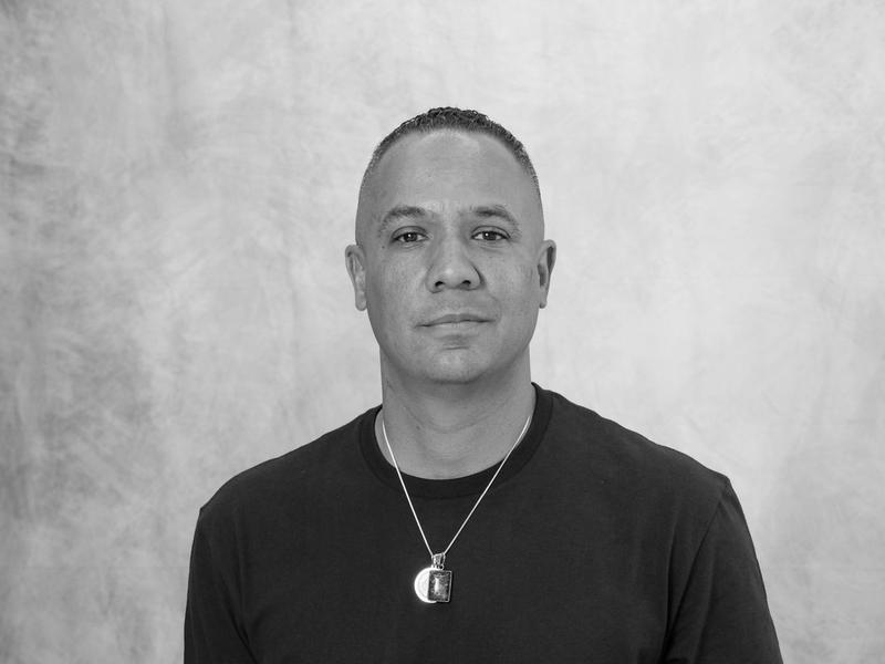 Black and white studio portrait of a man in a black tee against a painted canvas backdrop