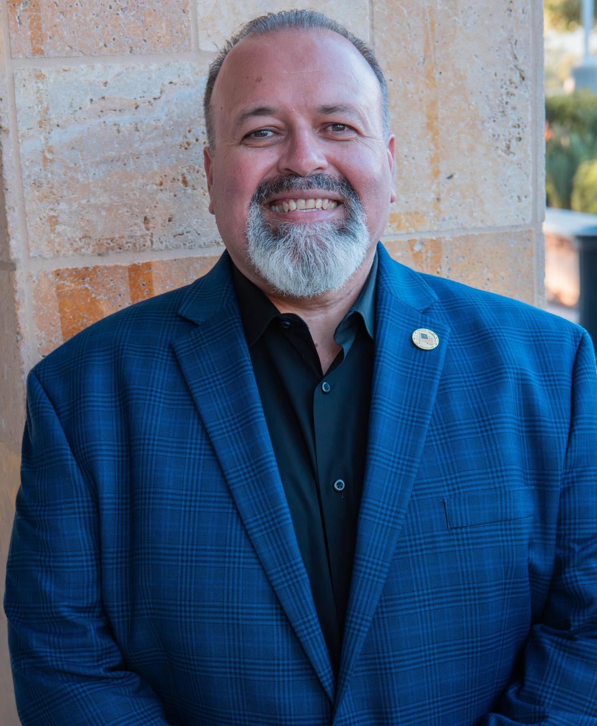 Warm outdoor portrait of a smiling man in a blue blazer against a sandstone wall at golden hour