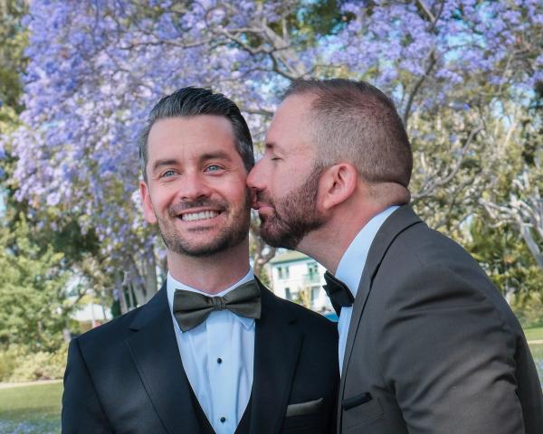 Groom kissing his partner on the cheek beneath the jacarandas
