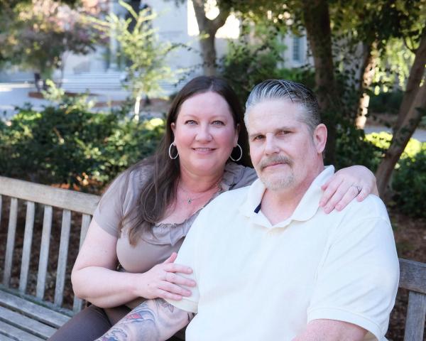 Older couple cuddled up on a wooden garden bench, smiling at the camera