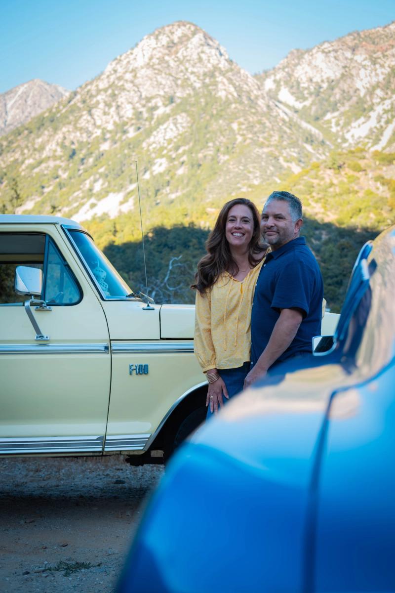 Couple hugging beside a cream Ford pickup with a mountain ridge behind them